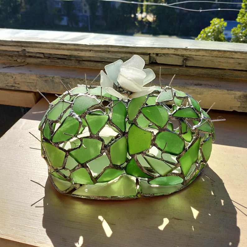 May include: A green glass cactus sculpture with a white glass flower on top. The sculpture is made of many small pieces of glass that are joined together with metal.