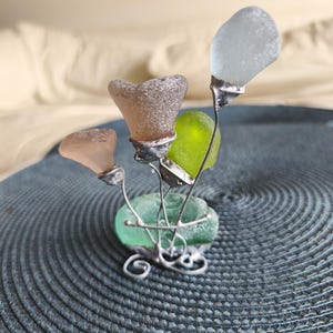May include: A decorative sculpture of sea glass flowers. The artwork features four flower-shaped pieces of sea glass in shades of pink, brown, green, and white, attached to silver wire stems. The base is a piece of green sea glass with decorative wire swirls.