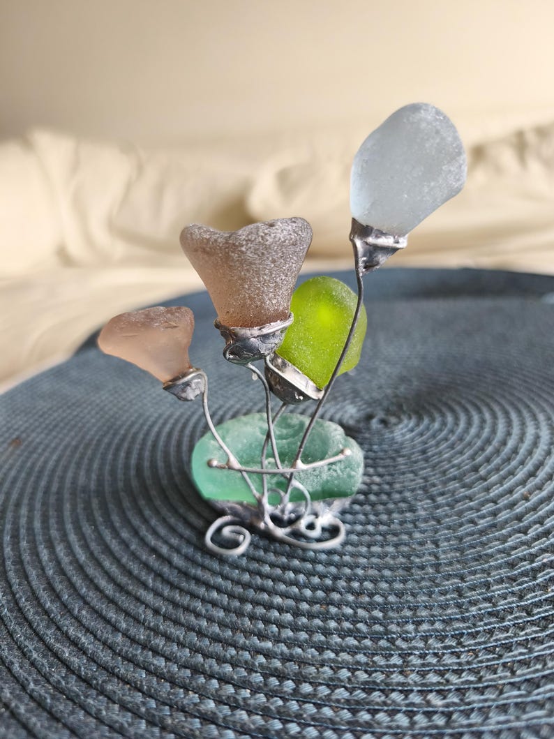May include: A decorative sculpture of sea glass flowers. The artwork features four flower-shaped pieces of sea glass in shades of pink, brown, green, and white, attached to silver wire stems. The base is a piece of green sea glass with decorative wire swirls.