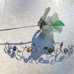 Op de afbeelding: Zeeglasbloem met een draadsteel en de woorden "Sea Stoned Glass" geschreven in draad met zeeglasaccenten.