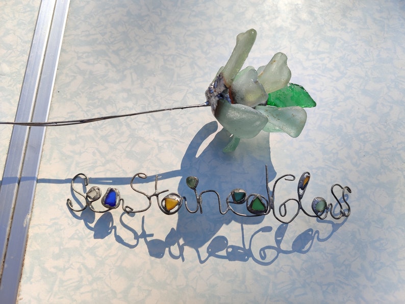 Op de afbeelding: Zeeglasbloem met een draadsteel en de woorden "Sea Stoned Glass" geschreven in draad met zeeglasaccenten.