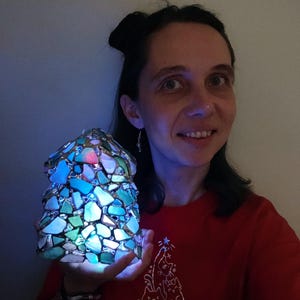 K&ouml;nnte beinhalten: Eine Person h&auml;lt einen blauen und gr&uuml;nen Weihnachtsbaum aus Glasmalerei mit einem Licht im Inneren. Der Baum besteht aus kleinen Glassplittern, die in einem Muster angeordnet sind. Die Person tr&auml;gt ein rotes T-Shirt mit einem Katzendesign.
