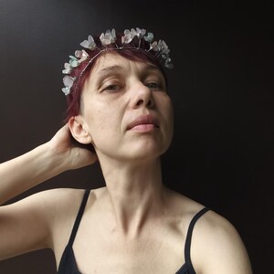 May include: A delicate silver-toned wire crown adorned with translucent, pale green and white flower-shaped beads. The crown is worn on red hair against a dark background. The person is wearing a black tank top.