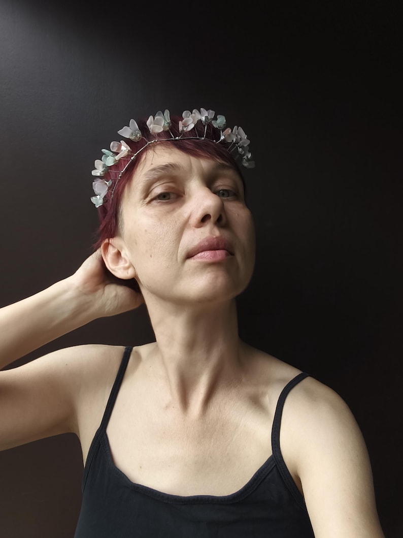 May include: A delicate silver-toned wire crown adorned with translucent, pale green and white flower-shaped beads. The crown is worn on red hair against a dark background. The person is wearing a black tank top.