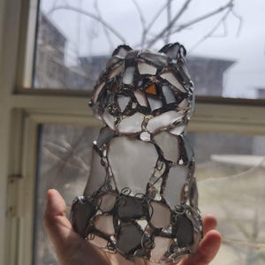 May include: A handcrafted cat figurine made of fused glass. The cat is constructed from various shapes of white, grey, and orange glass pieces, held together with a dark metal wire. The figurine is held in a hand, with a window and bare tree branches in the background.