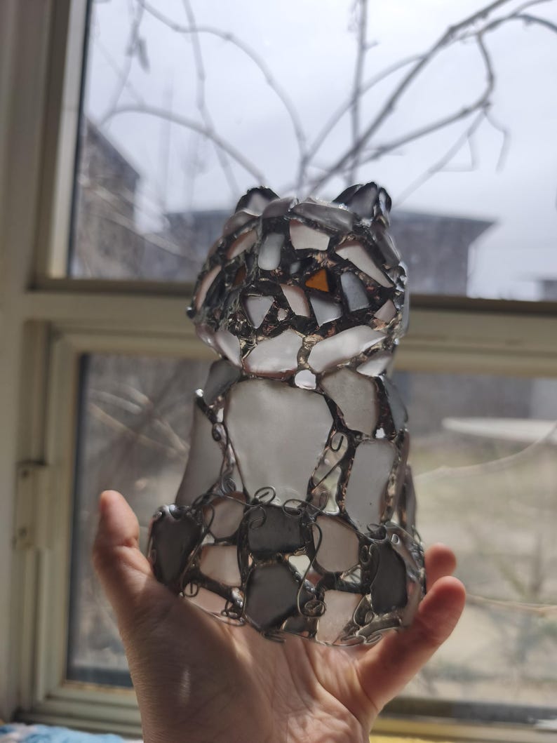 May include: A handcrafted cat figurine made of fused glass. The cat is constructed from various shapes of white, grey, and orange glass pieces, held together with a dark metal wire. The figurine is held in a hand, with a window and bare tree branches in the background.