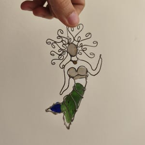 Puede incluir: Adorno de sirena hecho a mano con alambre y vidrio de colores. La sirena tiene un marco de alambre con una cara blanca, una cola verde y una aleta azul. El adorno se cuelga de un alambre fino.