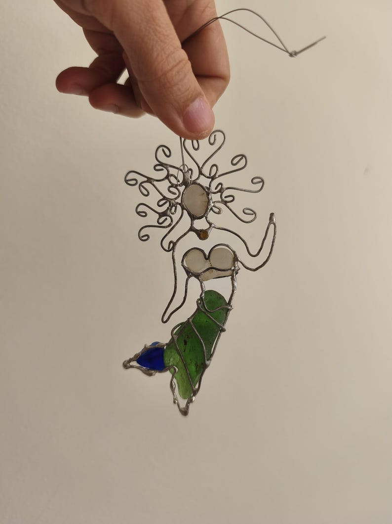 Puede incluir: Adorno de sirena hecho a mano con alambre y vidrio de colores. La sirena tiene un marco de alambre con una cara blanca, una cola verde y una aleta azul. El adorno se cuelga de un alambre fino.
