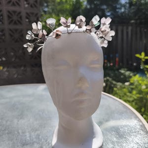 May include: A delicate floral crown with translucent, petal-shaped accents. The crown is made of thin, silver-colored wire and is displayed on a white mannequin head. The background features a blurred outdoor scene with greenery.