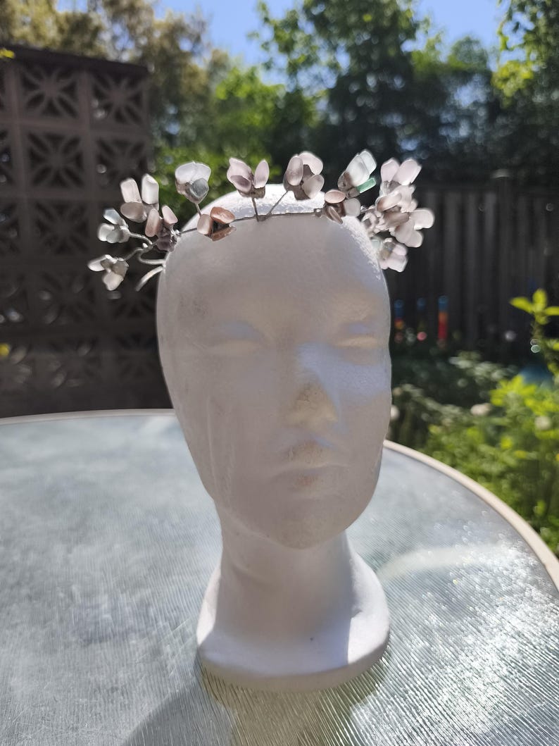 May include: A delicate floral crown with translucent, petal-shaped accents. The crown is made of thin, silver-colored wire and is displayed on a white mannequin head. The background features a blurred outdoor scene with greenery.