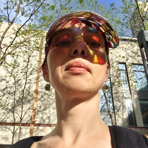 May include: A person wearing a colorful stained glass visor, with sunlight casting rainbow patterns across their face. The background includes trees, a building, and a blue sky. The image is taken from a low angle.