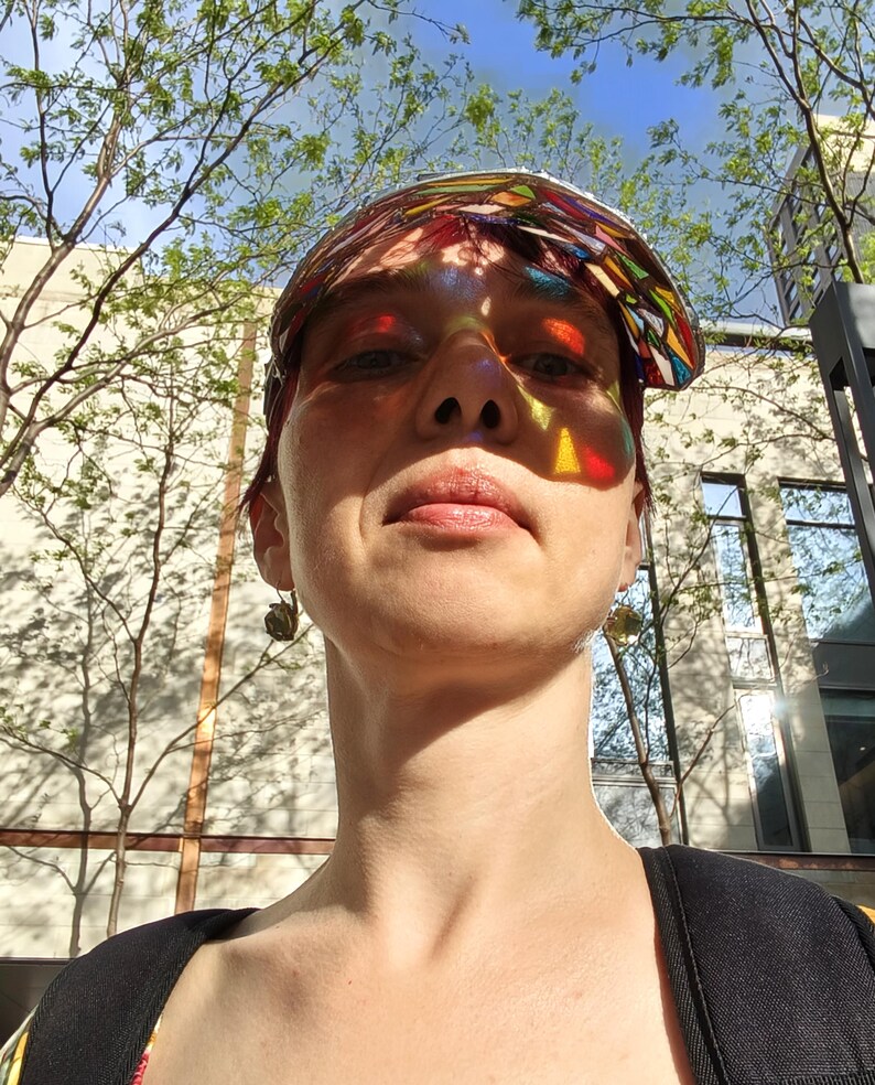 May include: A person wearing a colorful stained glass visor, with sunlight casting rainbow patterns across their face. The background includes trees, a building, and a blue sky. The image is taken from a low angle.