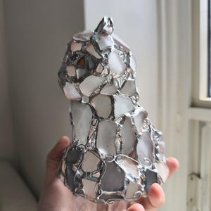 May include: A handmade cat sculpture made from sea glass and metal. The sea glass pieces are mostly white, with touches of pink and grey, and are held together by a silver-coloured metal frame. The sculpture is approximately 15 cm tall.