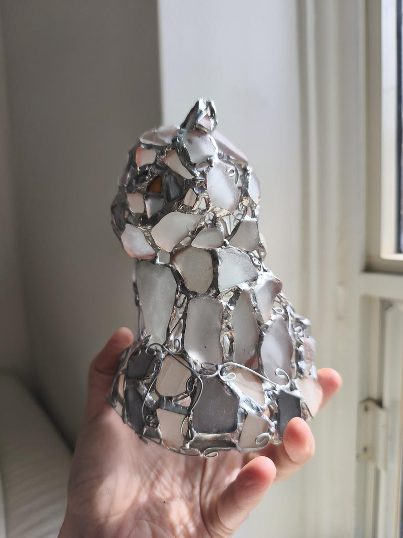 May include: A handmade cat sculpture made from sea glass and metal. The sea glass pieces are mostly white, with touches of pink and grey, and are held together by a silver-coloured metal frame. The sculpture is approximately 15 cm tall.