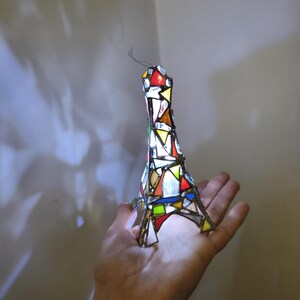 May include: A stained-glass Eiffel Tower ornament. The miniature tower is made from colourful glass pieces in red, yellow, green, and white, held together by metal. A wire extends from the top, giving it a delicate appearance.