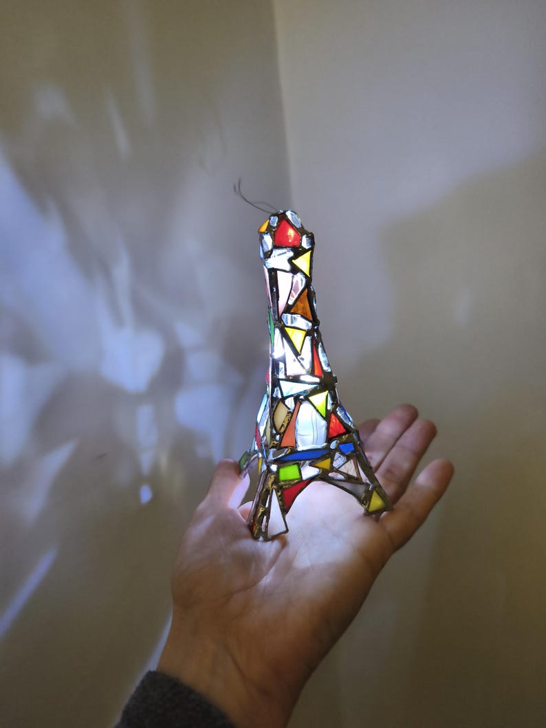 May include: A stained-glass Eiffel Tower ornament. The miniature tower is made from colourful glass pieces in red, yellow, green, and white, held together by metal. A wire extends from the top, giving it a delicate appearance.