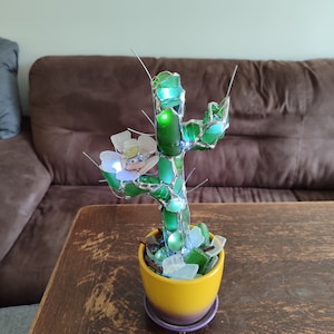 May include: A green glass cactus sculpture with a white flower, sitting in a yellow pot with a purple base. The cactus is made of many small pieces of green glass, and the flower is made of white and pink glass. The cactus is lit from within, and the light shines through the glass.