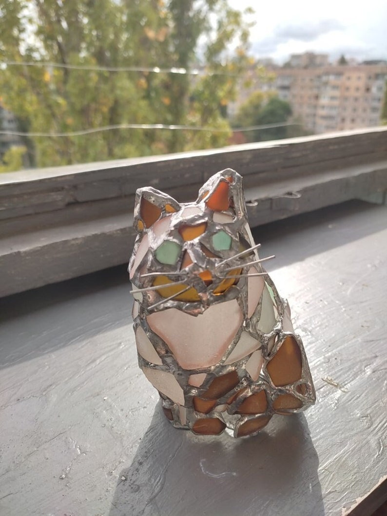 May include: A stained glass cat figurine with brown, white, and green glass pieces. The cat is sitting with its head tilted slightly to the side.