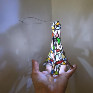 May include: A colourful stained glass ornament in the shape of a stylised tree, held in a hand. The ornament features a variety of colours including red, green, yellow, and blue. A wire loop is attached for hanging.