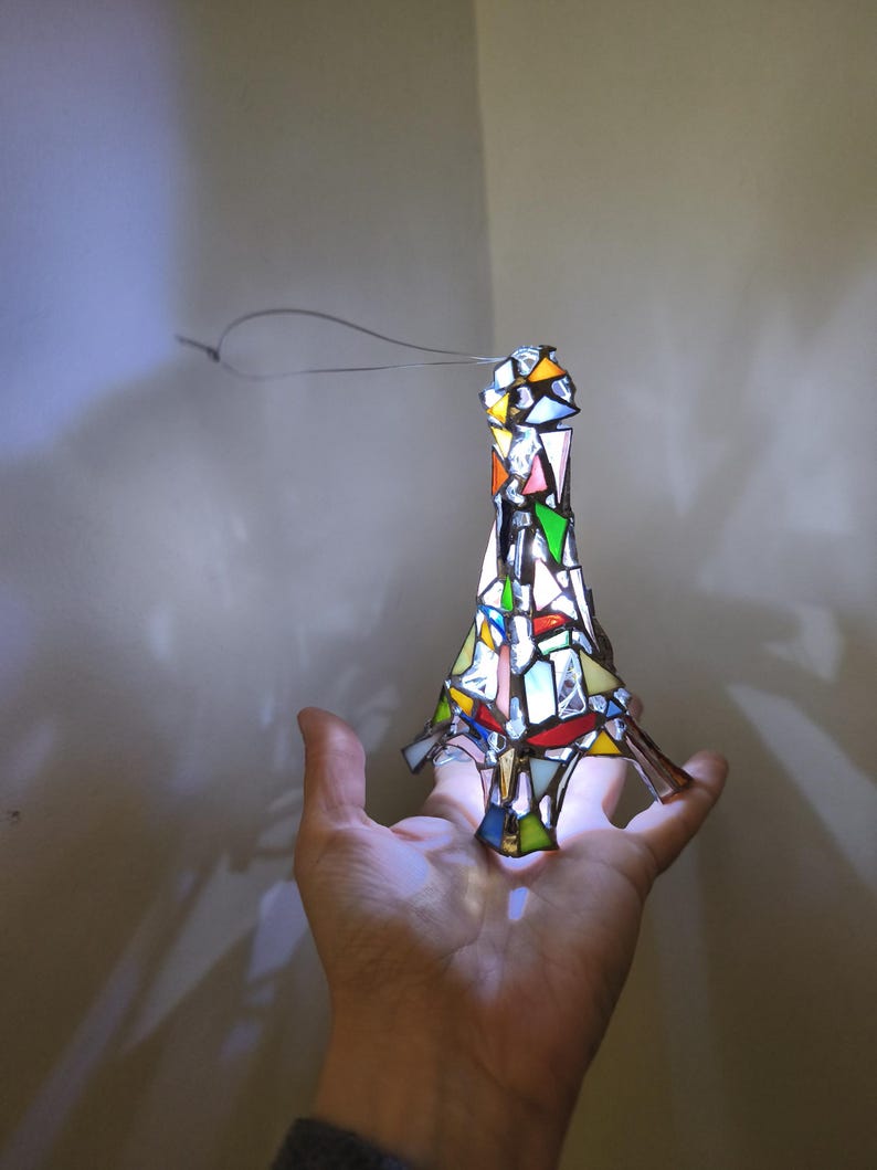 May include: A colourful stained glass ornament in the shape of a stylised tree, held in a hand. The ornament features a variety of colours including red, green, yellow, and blue. A wire loop is attached for hanging.