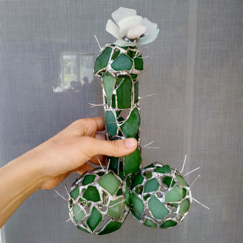 May include: A green glass sculpture of a cactus with a white flower on top. The cactus is made of many small pieces of green glass that are joined together.