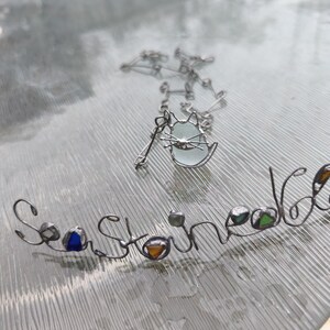 May include: A silver-toned necklace with a cat pendant and a separate wire-formed word, "Sea Stoned." The cat pendant has a light blue glass center. The word "Sea Stoned" has colorful glass accents.