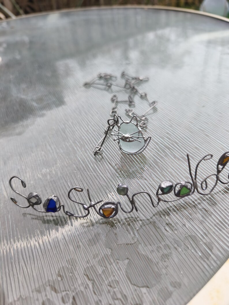 May include: A silver-toned necklace with a cat pendant and a separate wire-formed word, "Sea Stoned." The cat pendant has a light blue glass center. The word "Sea Stoned" has colorful glass accents.