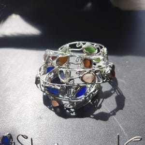 May include: Stack of three silver-toned wire bracelets, each adorned with colorful, irregularly shaped glass stones. The stones are in shades of green, orange, blue, and pink. The bracelets are stacked on a dark surface.