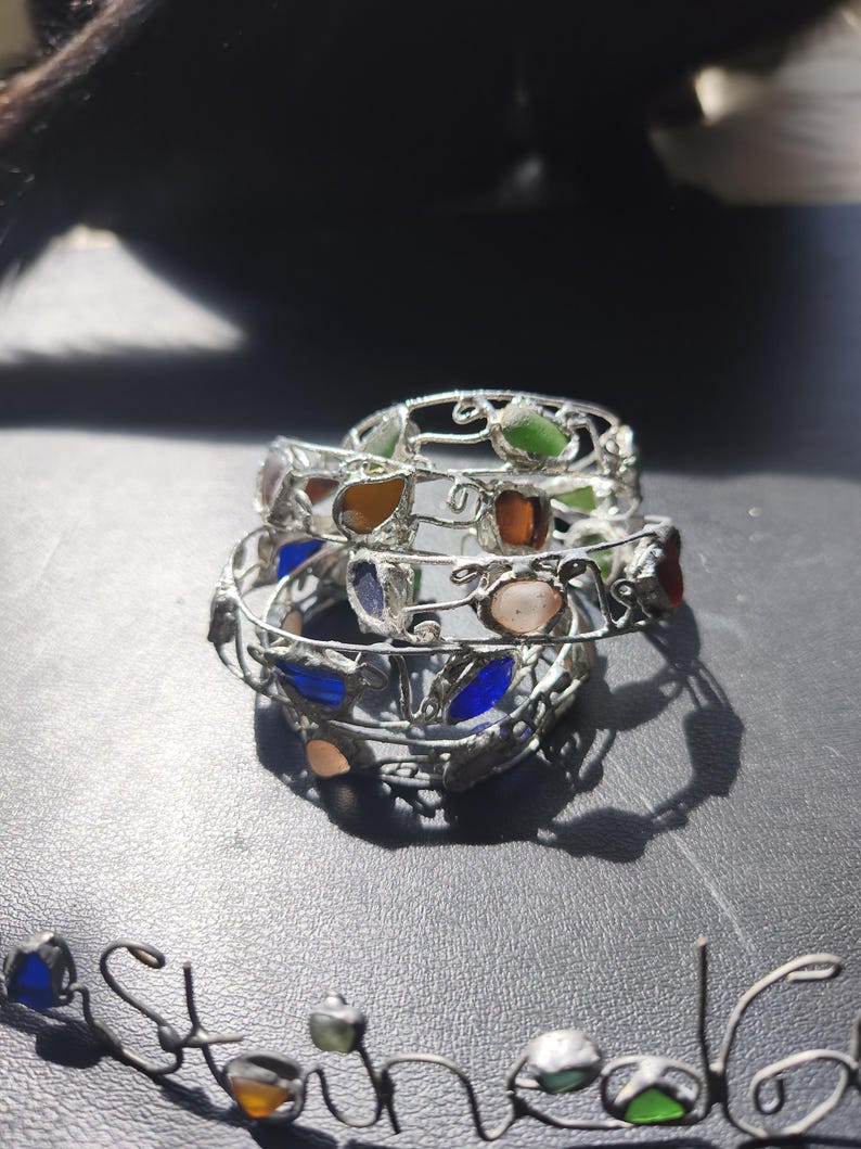 May include: Stack of three silver-toned wire bracelets, each adorned with colorful, irregularly shaped glass stones. The stones are in shades of green, orange, blue, and pink. The bracelets are stacked on a dark surface.