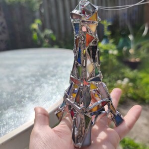 May include: A stained glass Eiffel Tower ornament. The tower is constructed from a mosaic of coloured glass pieces, including red, blue, yellow, and orange, joined by silver solder. The ornament is held in a hand against a blurred outdoor backdrop.