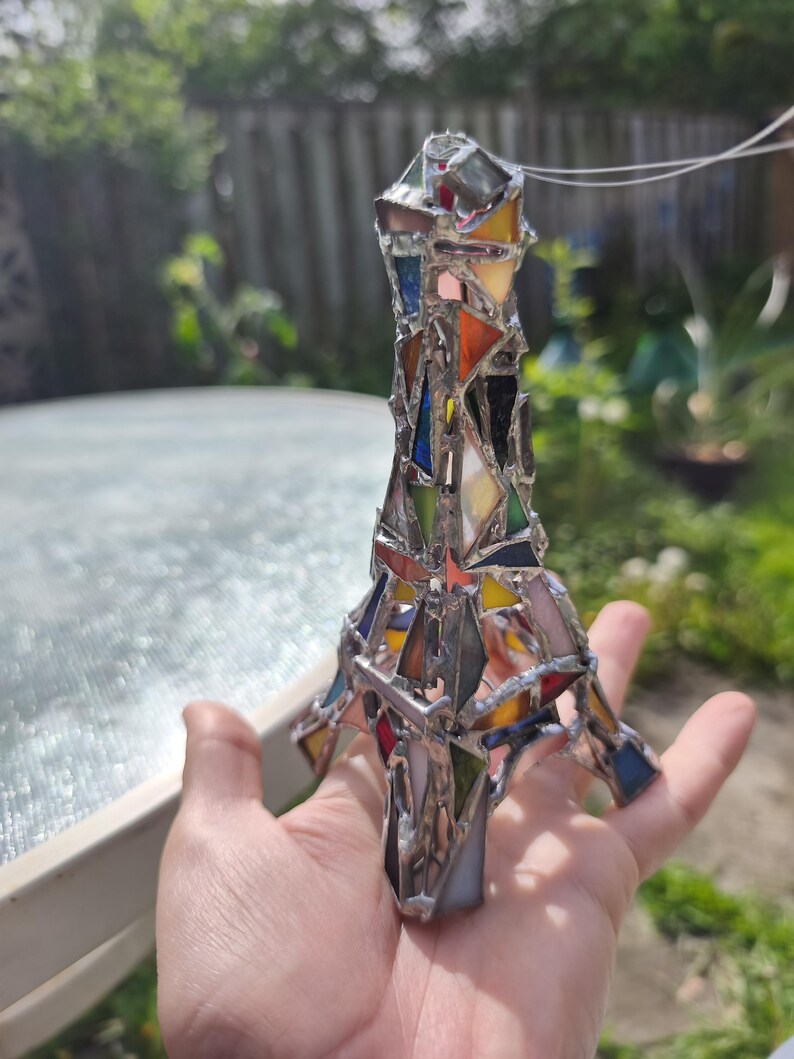 May include: A stained glass Eiffel Tower ornament. The tower is constructed from a mosaic of coloured glass pieces, including red, blue, yellow, and orange, joined by silver solder. The ornament is held in a hand against a blurred outdoor backdrop.