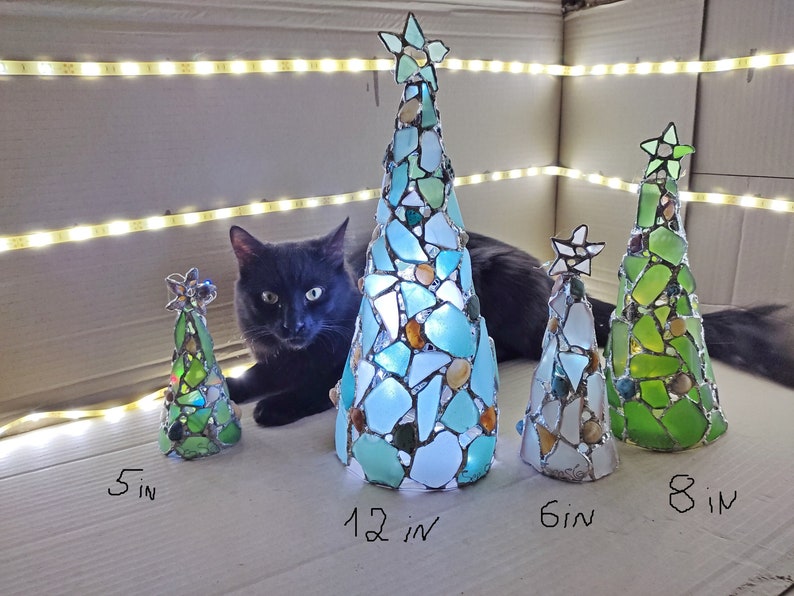 Pode incluir: Uma cole&ccedil;&atilde;o de &aacute;rvores de Natal de vidro feitas &agrave; m&atilde;o em v&aacute;rios tamanhos: 12, 15, 20 e 30 cm de altura. Cada &aacute;rvore tem formato c&ocirc;nico, adornada com pe&ccedil;as de vidro coloridas e encimada por uma estrela. As &aacute;rvores s&atilde;o iluminadas, criando um brilho festivo.