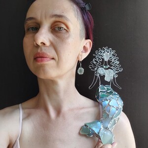 Puede incluir: Una figura decorativa de sirena hecha con alambre de tono plateado y trozos de vidrio marino en tonos azules y verdes. La sirena presenta una cabeza y torso formados por alambre con detalles de vidrio y una cola hecha de piezas de vidrio.