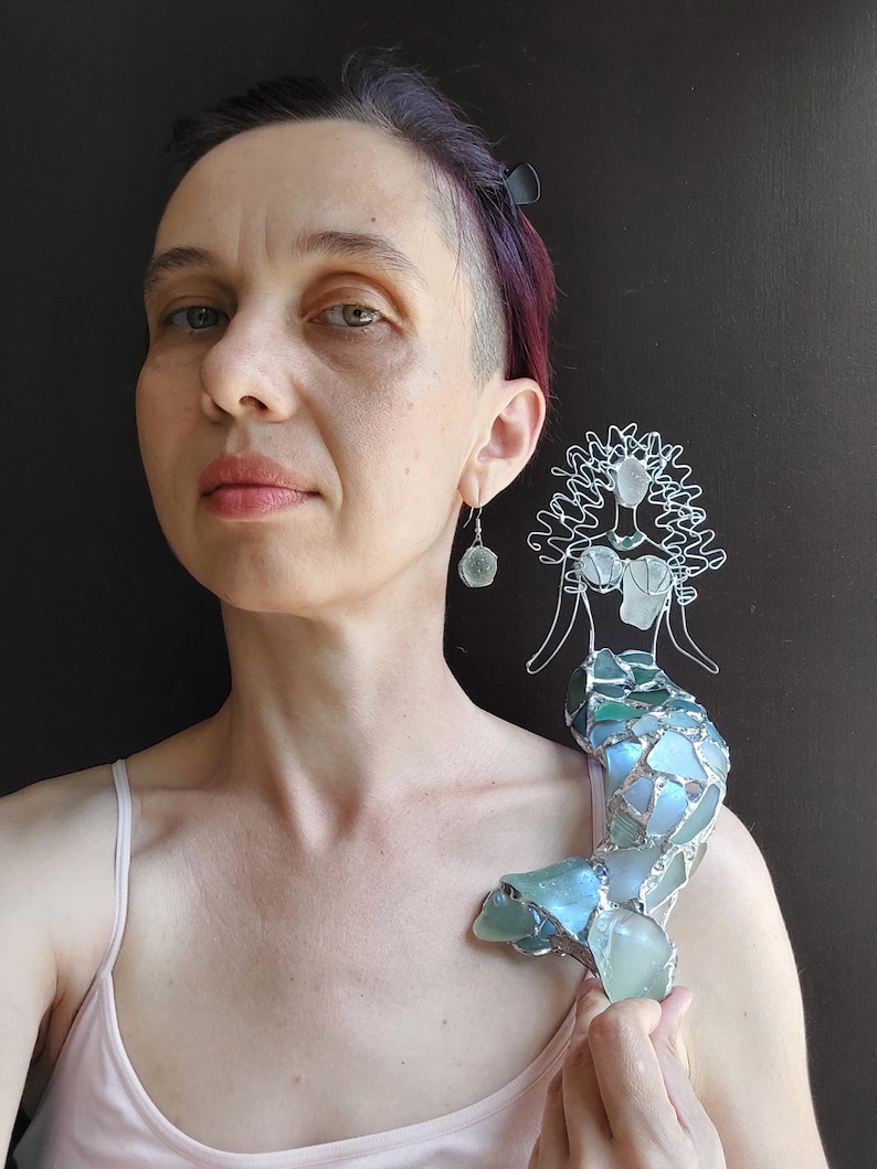 Puede incluir: Una figura decorativa de sirena hecha con alambre de tono plateado y trozos de vidrio marino en tonos azules y verdes. La sirena presenta una cabeza y torso formados por alambre con detalles de vidrio y una cola hecha de piezas de vidrio.