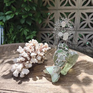 Puede incluir: Una escultura decorativa de sirena hecha de alambre de plata y piezas de vidrio marino verde transl&uacute;cido, con una cola y cabello detallados, colocada junto a una rama de coral blanco texturizada sobre una superficie de madera circular.