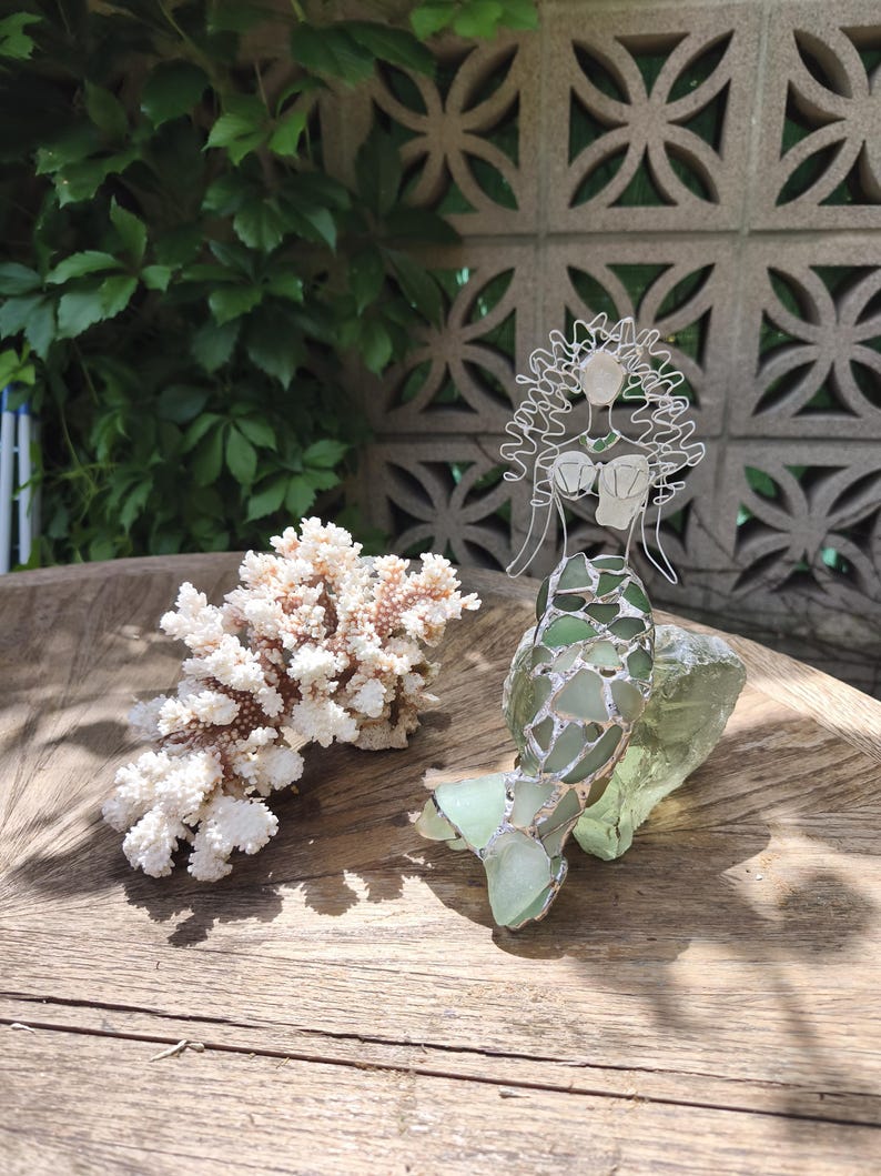Puede incluir: Una escultura decorativa de sirena hecha de alambre de plata y piezas de vidrio marino verde transl&uacute;cido, con una cola y cabello detallados, colocada junto a una rama de coral blanco texturizada sobre una superficie de madera circular.