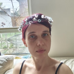 May include: A woman with short, red hair wearing a delicate floral crown. The crown features small, translucent, light blue and white flower-shaped beads. She is wearing a black tank top. The background includes a window and a white couch.