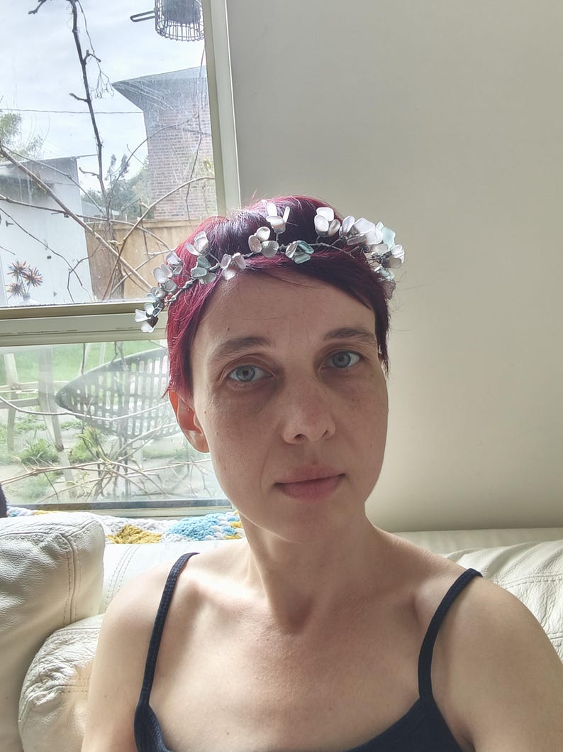 May include: A woman with short, red hair wearing a delicate floral crown. The crown features small, translucent, light blue and white flower-shaped beads. She is wearing a black tank top. The background includes a window and a white couch.