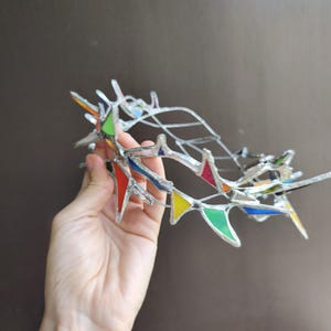 May include: A crown-shaped headpiece constructed from silver-toned metal wire and small, triangular stained glass pieces in vibrant colors including red, yellow, green, blue, and orange, arranged in a jagged, pointed, and open-work design.