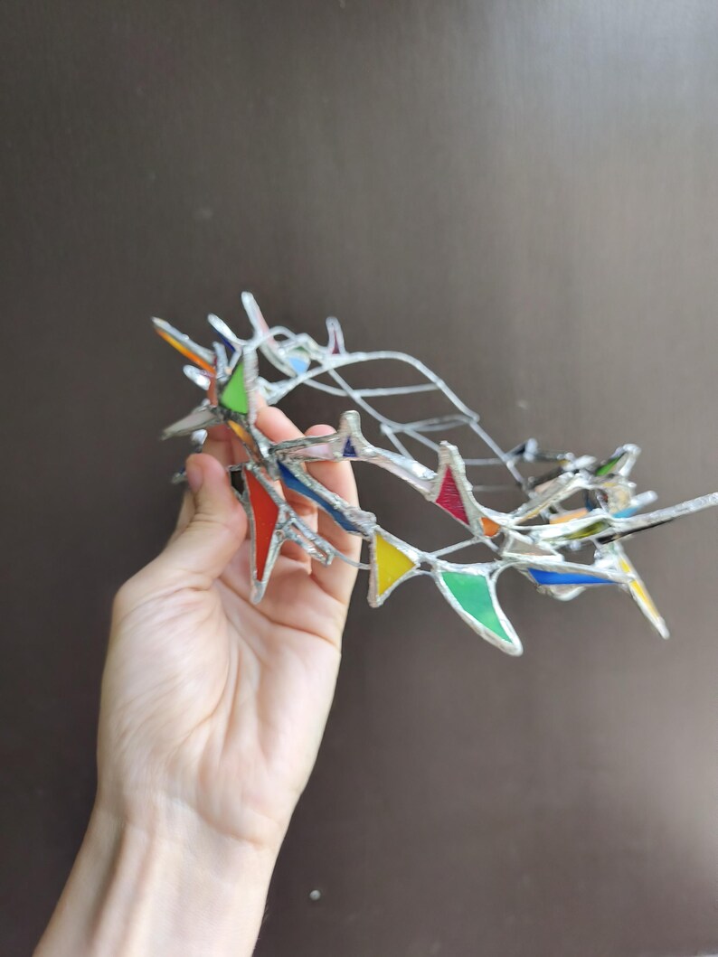 May include: A crown-shaped headpiece constructed from silver-toned metal wire and small, triangular stained glass pieces in vibrant colors including red, yellow, green, blue, and orange, arranged in a jagged, pointed, and open-work design.