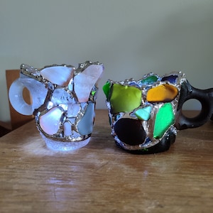 May include: Two small ceramic mugs, one white with light blue and pink accents, the other black with green, yellow, and orange accents. Both mugs are made of mosaic pieces of glass.