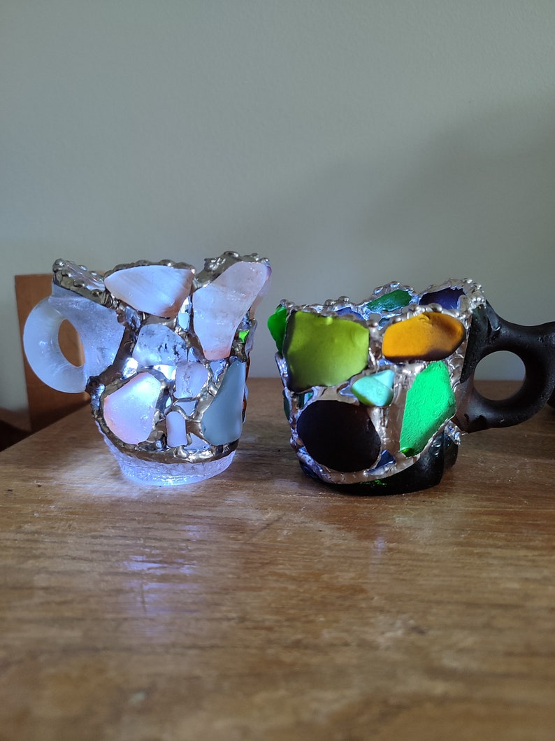 May include: Two small ceramic mugs, one white with light blue and pink accents, the other black with green, yellow, and orange accents. Both mugs are made of mosaic pieces of glass.