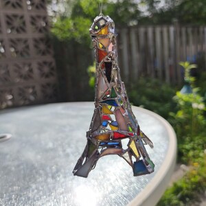 May include: A stained glass Eiffel Tower ornament in a patchwork design. The miniature tower is made of various coloured glass pieces, including red, yellow, blue, and green, held together by silver solder. The ornament is suspended by a thin wire.