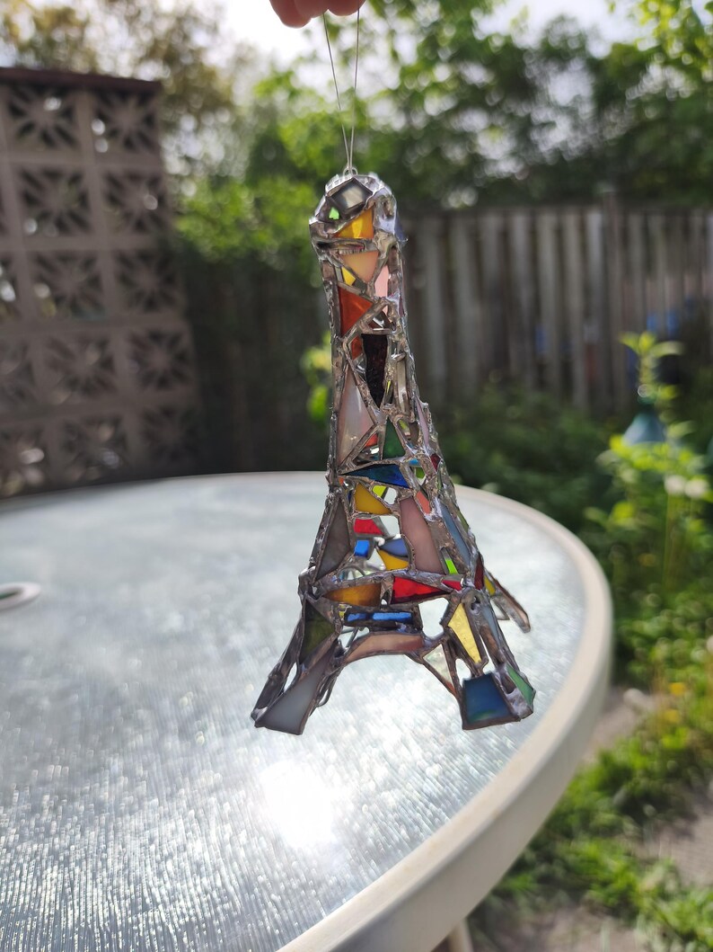 May include: A stained glass Eiffel Tower ornament in a patchwork design. The miniature tower is made of various coloured glass pieces, including red, yellow, blue, and green, held together by silver solder. The ornament is suspended by a thin wire.