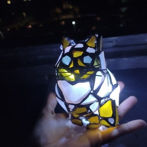 May include: A stained glass cat figurine with a white, yellow, and black design. The cat is illuminated from within, creating a warm glow.