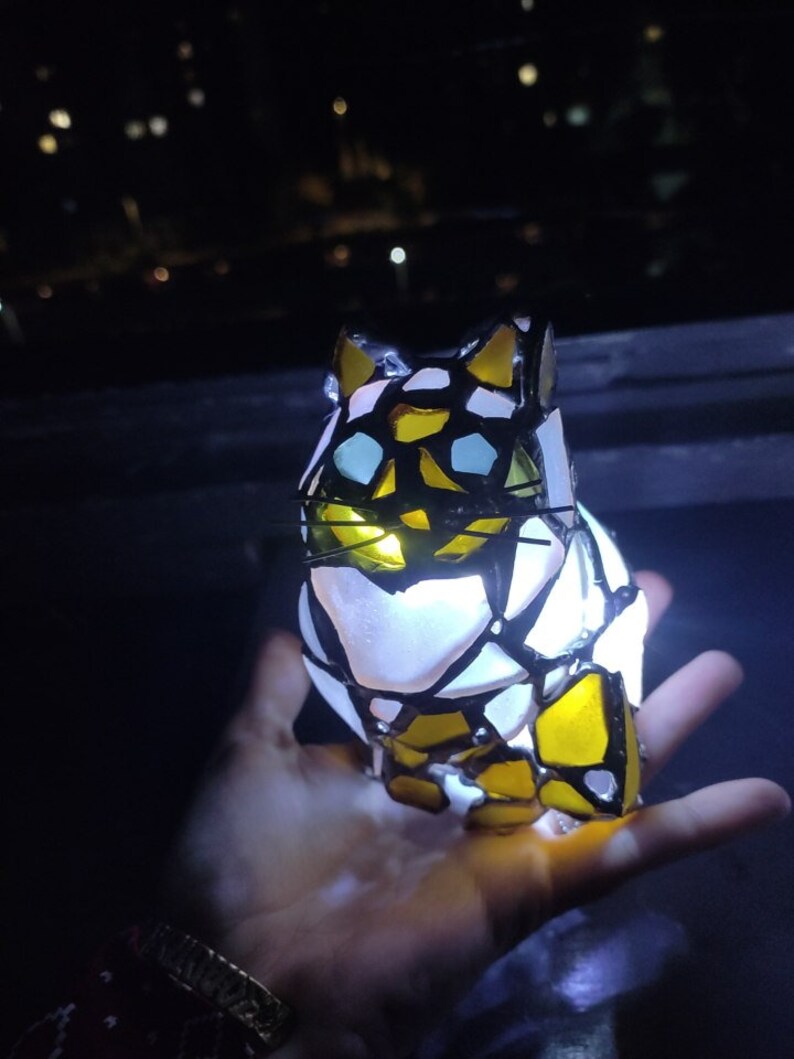 May include: A stained glass cat figurine with a white, yellow, and black design. The cat is illuminated from within, creating a warm glow.