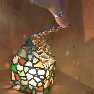 May include: A stained glass candle holder shaped like a house with a removable roof. The house is made of small pieces of glass in shades of green, brown, and white. The candle holder is lit and the light shines through the glass.