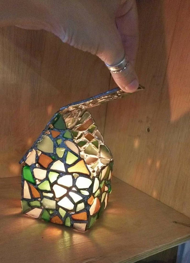 May include: A stained glass candle holder shaped like a house with a removable roof. The house is made of small pieces of glass in shades of green, brown, and white. The candle holder is lit and the light shines through the glass.
