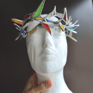 May include: A crown made of stained glass pieces in red, yellow, green, and blue, held together by silver-toned metal solder. The crown features sharp, pointed spikes of varying heights and is displayed on a white, featureless mannequin head.