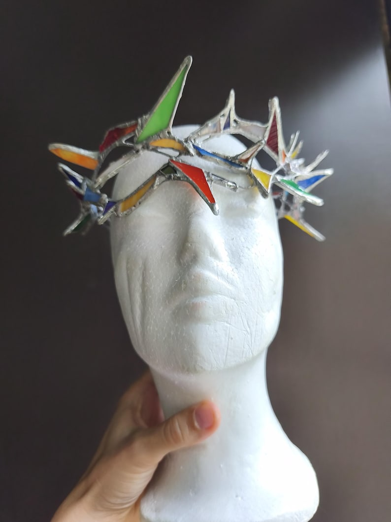 May include: A crown made of stained glass pieces in red, yellow, green, and blue, held together by silver-toned metal solder. The crown features sharp, pointed spikes of varying heights and is displayed on a white, featureless mannequin head.
